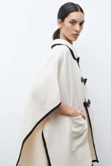 Capa color crudo con cuello camisero y ribetes negros. Cierre frontal con alamares negros y bolsillos laterales.