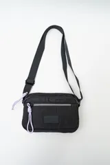 Cartera tipo bandolera de nylon color negro, con cierre frontal y detalles de tiradores en color lila. Cuenta con correa ajustable y parche con logo en el frente.