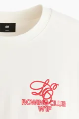 Remera boxy de algodón color blanco, con cuello redondo acanalado, mangas cortas y hombros caídos. Presenta un pequeño estampado rojo en el pecho con las inscripciones 'ROWING CLUB W1F'.