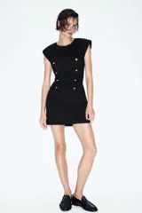 Vestido corto de estilo militar en color negro, con cuello redondo, hombreras y manga sisa. Presenta un diseño frontal con falsos botones dorados dispuestos en filas horizontales. Cierre lateral con cremallera oculta y abertura en la espalda con botón.