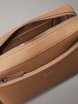Bandolera Calvin Klein color tostado, con correa ancha de tela con logo de la marca y correa fina de cuero sintético.