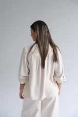 Blusa de lino color beige, con cuello en V, cierre frontal con botones y detalle de alforzas y encaje en el pecho. Presenta mangas largas con puños sencillos y un corte holgado.