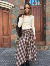 Pollera midi de corte amplio con estampado escocés en tonos marrones. Presenta un diseño distintivo con un detalle de atado en la cintura que simula puños de camisa y cierre frontal con botones.