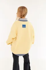 Sweater de punto color amarillo con silueta oversize y estilo universitario vintage. Presenta cuello redondo, puños y ruedo en tejido acanalado de color gris azulado, y un diseño gráfico bordado en el frente.