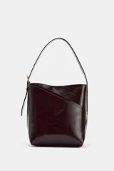 Bolso tipo saca de diseño asimétrico con acabado brillante, asa ajustable con hebilla lateral y cierre mediante botón imán.