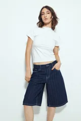 Bermuda de denim en color azul oscuro con diseño de rayas verticales blancas. Presenta un corte holgado, tiro medio y cierre frontal con botón y cremallera.