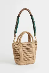 Cartera Rapsodia tipo tote mini en color tostado, confeccionada con cuerpo de tejido estilo red y detalles en material sintético liso. Cuenta con asas de mano acolchadas y una correa desmontable trenzada en tonos verde, rosa y marrón con herrajes metálicos. Incluye una placa frontal con el nombre de la marca grabado.