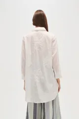Camisa blanca de manga larga, corte holgado y largo, con cuello camisero y botones frontales. Presenta bordados florales en tono blanco sobre el tejido.