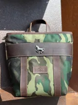 Mochila pequeña Sierra Mora con diseño de camuflaje, confeccionada en tela resistente con detalles en cuero marrón. Presenta un aplique metálico en forma de caballo en el frente y una etiqueta de cuero con el logo de la marca.
