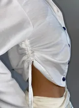 Camisa blanca de corte cropped con ajuste mediante cordones laterales y botones en contraste de color azul oscuro. Presenta cuello clásico, mangas largas con puños y diseño ligero.