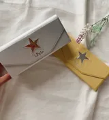 Estuche para lentes plegable con diseño rígido y cierre magnético. Presenta una estrella metálica decorativa en la solapa frontal y el logo de la marca estampado. El interior está forrado en material suave para proteger los cristales.