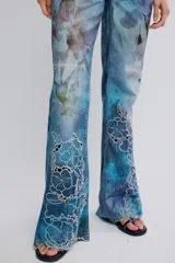 Jean de tiro medio y pierna flare de denim azul con estampa en toda la prenda y detalles de calados bordados florales en la parte inferior. Cuenta con cinco bolsillos y cierre frontal con cremallera.