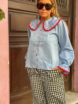 Camisa de color celeste con cuello bobo grande y puños con volados, ambos con ribetes en color rojo. Presenta cierre frontal con botones rojos.