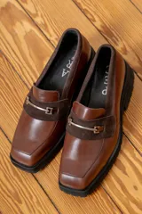 Mocasín de cuero vacuno color marrón, con suela track de goma negra, puntera cuadrada y detalle de herraje metálico sobre una banda de gamuza en el empeine.
