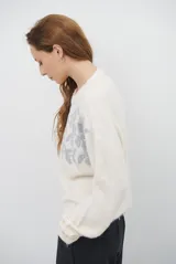 Buzo de tejido de punto en color beige, con cuello redondo y mangas largas. Presenta un delicado bordado de ramas y hojas en color crema en la parte superior del frente.