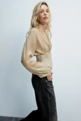 Top beige de manga larga con escote en pico, mangas abullonadas y puños elásticos. Presenta un diseño combinado con tejido fluido en la parte superior y tejido estructurado tipo corsé en la cintura, con cierre frontal de ganchos metálicos.
