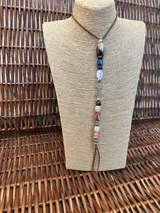 Colgante largo de estilo artesanal con cordón de cuero marrón y una serie de cuentas de piedra natural en diversos colores y texturas, intercaladas con elementos metálicos decorativos.