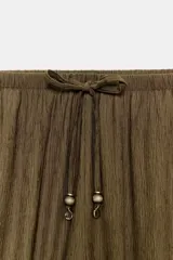 Pantalón fluido de tiro alto color verde militar, con cintura elástica ajustable con cordones y detalles de cuentas de madera. Tiene bolsillos delanteros y pierna recta.