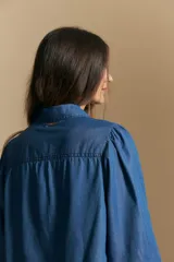 Camisa de denim azul con cuello camisero, cierre frontal de botones y mangas largas abullonadas con hombros fruncidos.