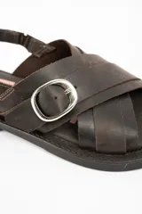 Sandalias de cuero marrón oscuro, con tiras cruzadas en el empeine, hebilla metálica grande decorativa y tira ajustable en el talón con hebilla plateada.