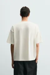 Camiseta de corte relajado color crudo, con cuello redondo y manga corta. Presenta un estampado gráfico en el frente con motivos de palmeras y paisajes en color negro.