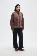 Campera de diseño híbrido que combina paneles de corderito sintético en color crema con mangas y bolsillos de tela técnica acolchada en color verde militar. Presenta cierre frontal completo, cuello alto y un pequeño parche decorativo en el pecho.