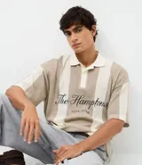 Remera polo de punto con diseño de rayas verticales en tonos beige y blanco. Presenta un corte oversized, cuello clásico con abertura de botones y bordado frontal con la inscripción The Hamptons.