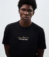 Remera de algodón color negro con cuello redondo y manga corta. Presenta un pequeño estampado gráfico en el centro del pecho con la frase 'Wild and Free'.