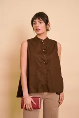 Blusa sin mangas de color blanco, con cuello mao y cierre frontal de botones. Presenta un diseño holgado y asimétrico, con la parte trasera más larga y aberturas laterales en el dobladillo.