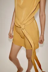 Vestido corto color mostaza con cuello fluido, manga sisa y hombreras marcadas. Presenta un detalle de lazada en la cintura y cierre de cremallera oculto en la espalda.
