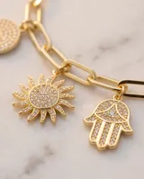 Pulsera de cadena de eslabones dorada con cinco dijes colgantes decorados con circonias: un corazón, una mano de Hamsa, un sol, un hueso de la suerte y un círculo.