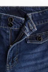 Jean de tiro alto con corte acampanado desde la rodilla. Presenta cierre frontal con doble botón, bolsillos frontales decorativos con solapa y botón, y bolsillos traseros tipo parche.