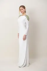 Vestido largo blanco de manga larga, con escote redondo al frente, espalda baja descubierta y detalles de bordado de piedras y pedrería en tonos verdes sobre los hombros.