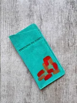 Portalentes de cuero gamuzado color turquesa, de diseño rectangular, con arte pintado a mano en tonos cobre y rojo. Presenta el logo de la marca grabado en la parte frontal.