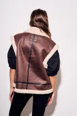 Chaleco oversize estilo gamulán en color marrón, confeccionado en cuero vegano con forro y solapas de corderito sintético en contraste. Posee cierre frontal metálico, bolsillos con cremallera y detalles de correas con hebillas en el cuello.