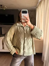Saco tejido en color verde oliva con diseño de ochos y trenzas. Presenta cuello estilo camisero, cierre frontal con botones tipo joya, mangas holgadas y silueta oversize.