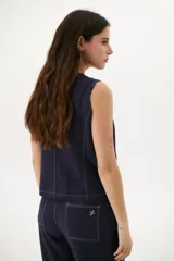 Chaleco sin mangas color crudo, con escote en V y cierre frontal de tres botones oscuros. Presenta costuras expuestas en contraste oscuro que definen el diseño y bolsillos delanteros simulados.