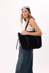 Bolso tipo tote de gamuza sintética en color marrón, con diseño amplio y manijas largas. Presenta dos líneas verticales de tachas metálicas plateadas en el frente y en los laterales.