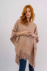 Ruana artesanal tejida en telar manual, con textura rústica y terminación en flecos. Presenta un diseño liviano en tonos rosados y naranjas, con etiqueta de marca en el frente.