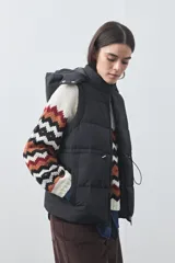 Chaleco puffer negro con diseño acolchado, capucha desmontable y cierre frontal. Cuenta con cordones ajustables en la cintura y bolsillos laterales.