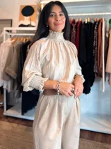 Blusa de tela liviana con diseño de rayas verticales en tonos beige y crema. Presenta cuello alto con detalle de fruncido tipo smock, mangas largas abullonadas con puños elásticos y volados en los extremos.