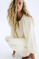 Blusa de manga globo larga color crema, con cuello redondo fruncido y botones ocultos en la parte delantera. Presenta un bajo redondeado.
