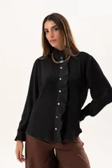 Camisa negra de textura sutil, con cuello alto tipo mao, cierre frontal con botones y detalle de volado lateral. Presenta mangas largas con puños con volados.