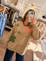Buzo de cuello redondo en color beige, con diseño oversize y felpa interna. Presenta bordados de flores en tonos rosa, verde y azul distribuidos en el frente.