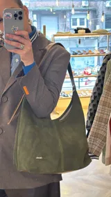 Cartera tipo hobo de gamuza verde oscuro, con textura grabada y costuras pespunteadas a la vista. Cuenta con una correa de hombro ajustable de cuero sintético marrón, detalles metálicos dorados y la marca 'chérie' grabada en la parte inferior frontal.