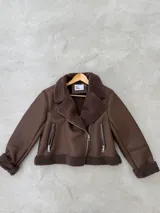 Campera tipo gamulán de cuero sintético en color marrón chocolate, con interior y solapas de corderito sintético al tono. Presenta cierre frontal cruzado y bolsillos laterales con cierre metálico.