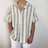 Camisa de manga corta con corte oversize, cuello tipo cubano y cierre frontal con botones. Presenta un diseño de rayas verticales en tonos crudo y gris.