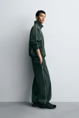 Pantalón jogger de corte relajado confeccionado en tejido técnico de color verde oscuro. Presenta cintura elástica con cordón interior, bolsillos laterales con cierre y bajo ajustable con elástico. Incluye detalles de líneas en contraste a lo largo de las piernas.