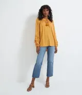 Blusa de manga larga color mostaza con textura de puntos en relieve (dobby). Presenta cuello redondo con abertura en V y cordón ajustable con borla (tassel) en el escote. Las mangas tienen detalles de entretejido calado (entremedios) y volados con terminación fruncida en los puños.