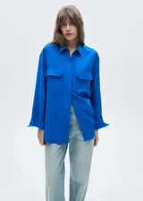 Camisa de corte recto y diseño largo, confeccionada en tejido fluido mezcla de lyocell y lino. Presenta cuello camisero, cierre frontal de botones ocultos bajo tapeta, mangas largas con puños abotonados y dos bolsillos de solapa en el frente.
